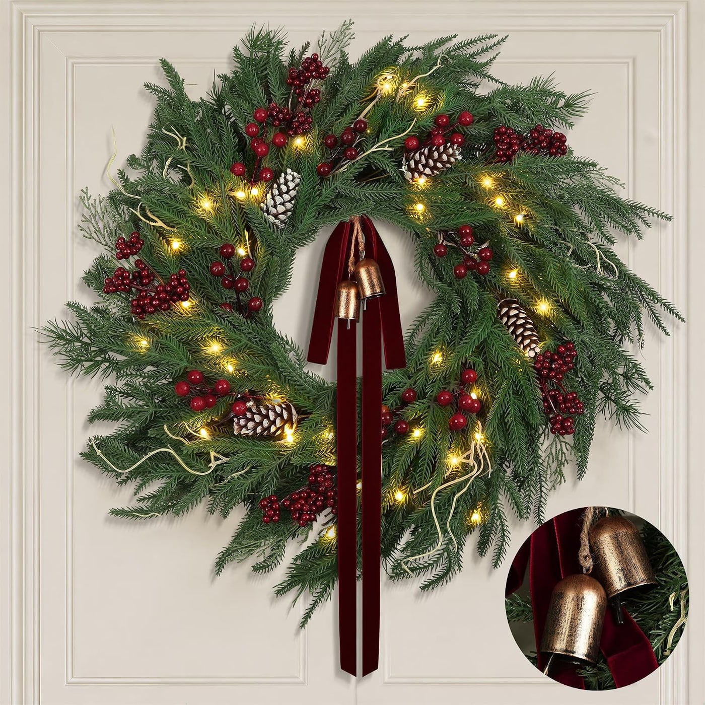 Lush faux Norfolk pine wreath with dense green branches, wine-red velvet bow, and gold bells